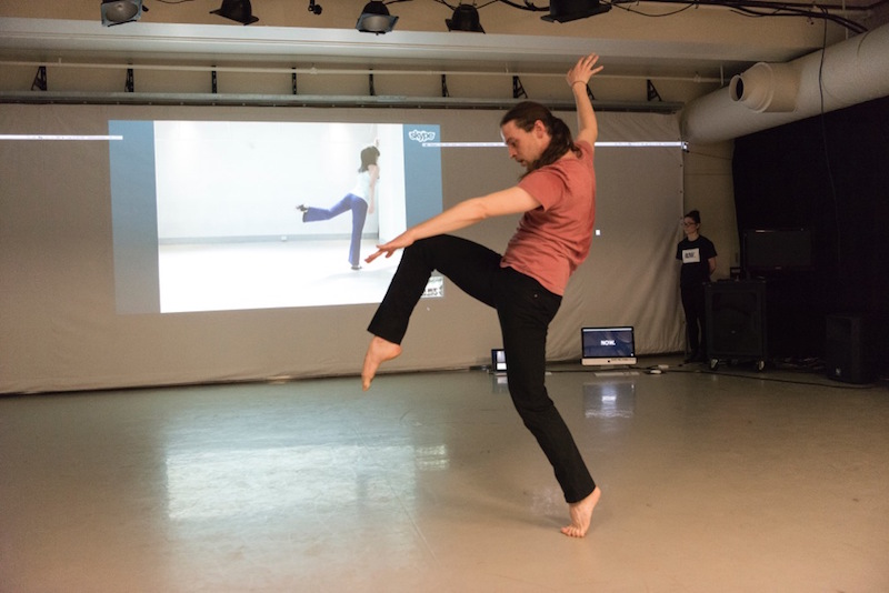 A man balances on one leg in the studio with his other leg hovering in front of him. His counterpart on video also balances on one leg but her other leg is behind her